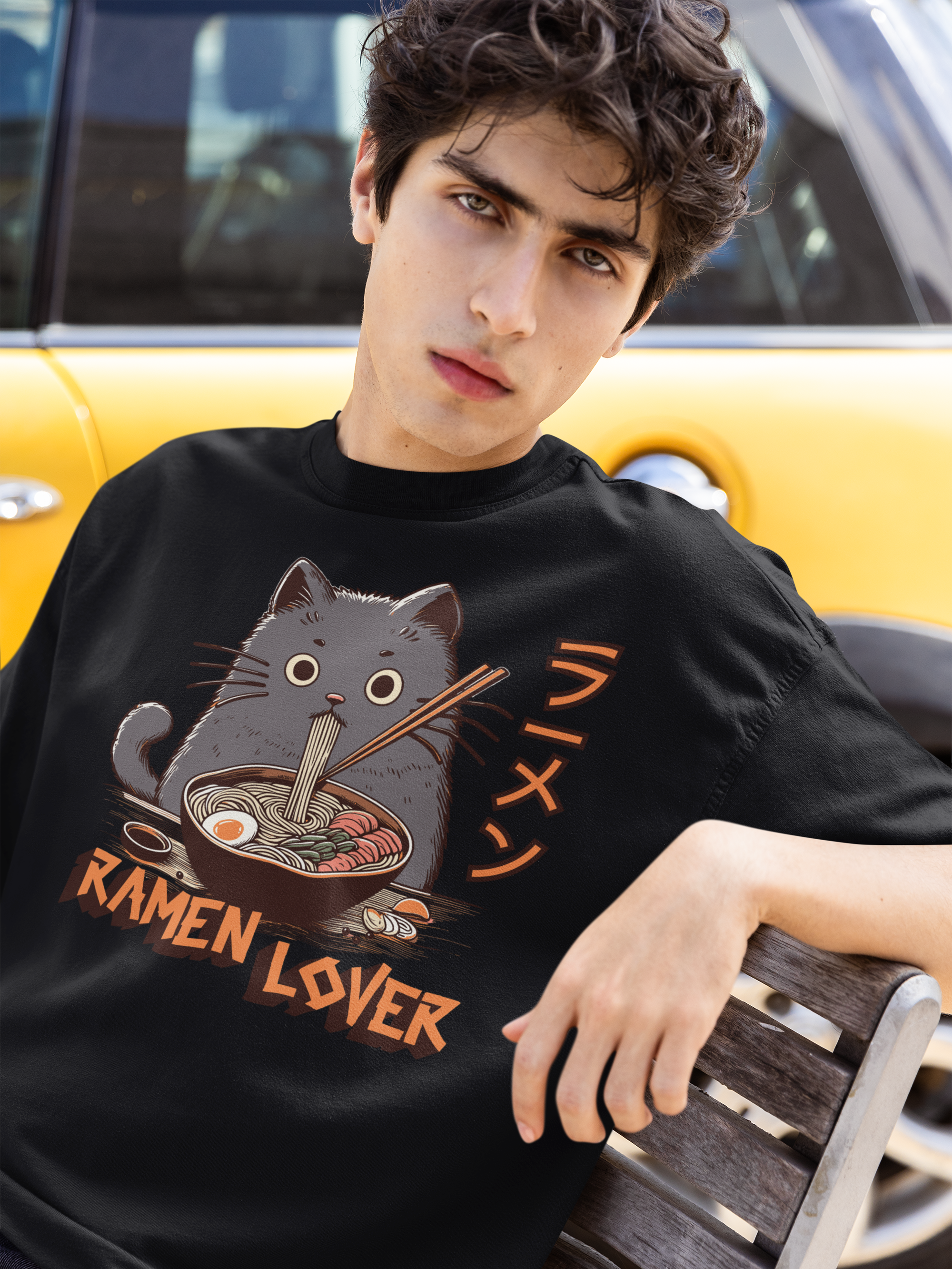 Person wearing a black t-shirt with a cat eating ramen graphic and 'Ramen Lover' text, sitting in front of a yellow car.